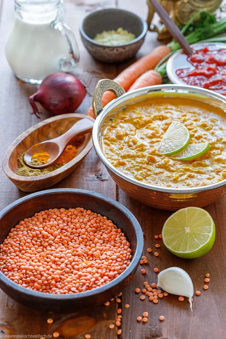 Alle frischen Zutaten für das rote Linsen Rezept: Rote Linsen, Karotten, Zwiebeln, Knoblauch, Ingwer, Kokosmilch, Tomaten und Gewürze übersichtlich auf einem Brett.