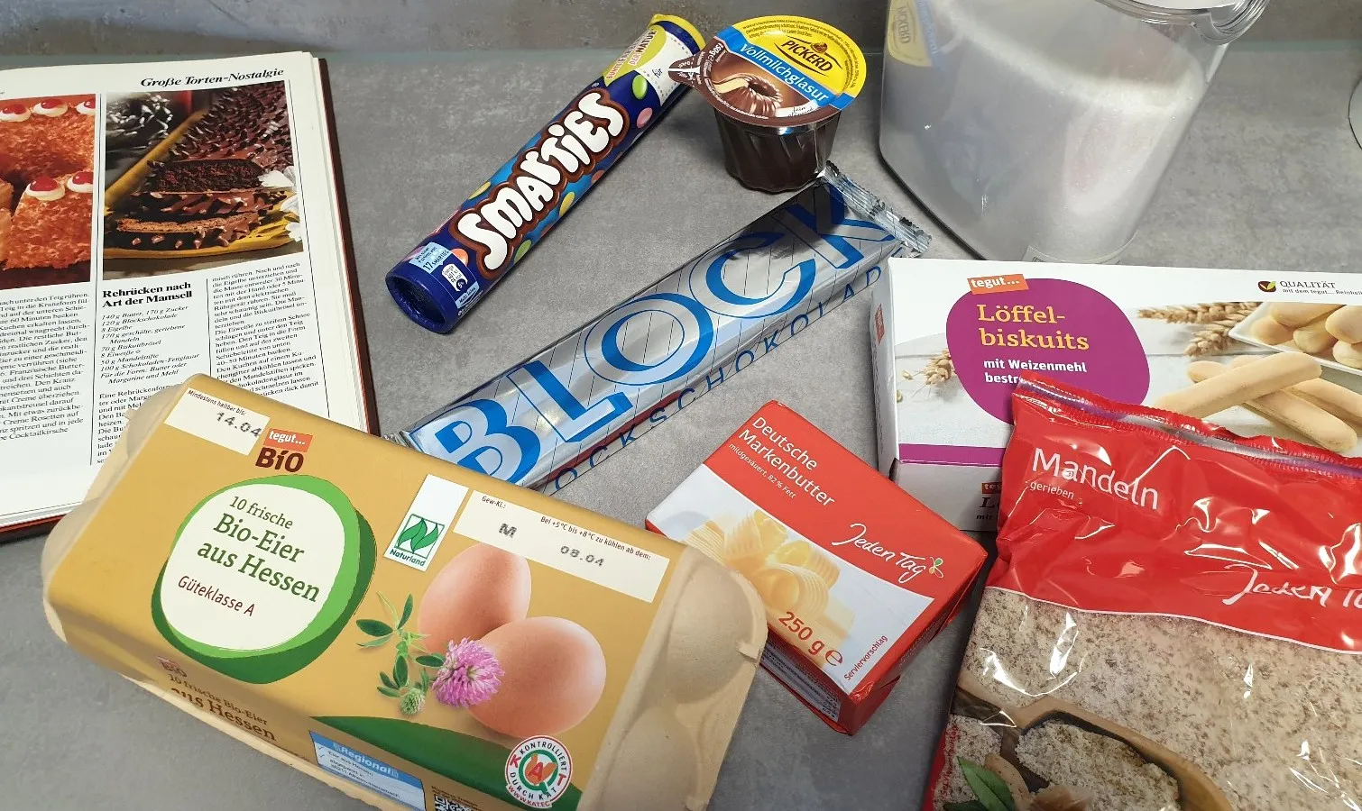 Alle Zutaten für den großen Geburtstagskuchen bereitgelegt: Butter, Zucker, Schokolade, Eier, Mandeln und Löffelbiskuit