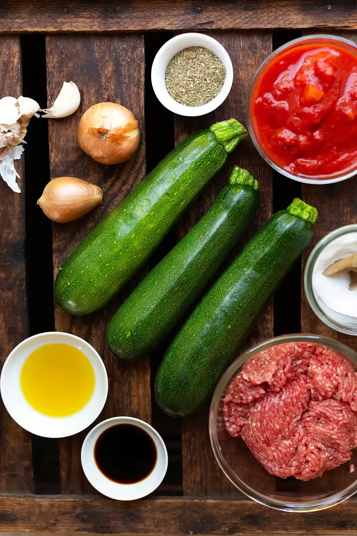 Alle Zutaten für die Zucchini-Hackfleisch-Pfanne in einzelnen Schälchen oder direkt auf einem dunklen Holzuntergrund. Zu sehen sind drei Zucchini, gehackte Tomaten, Hackfleisch, Zwiebel, Knoblauch, Öl, Sojasauce und Gewürze. Die frischen Zutaten sorgen für einen guten Geschmack.