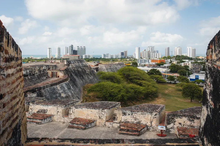 Alte Steinmauer mit Blick auf eine Grünfläche, im Hintergrund moderne Gebäude unter teilweise bewölktem Himmel