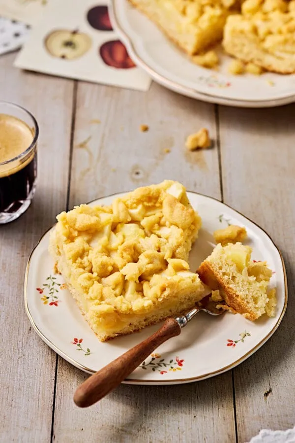 Apfelkuchen mit Hefeteig und Streusel auf einem Kuchenteller und einem Kaffee im Hintergrund.