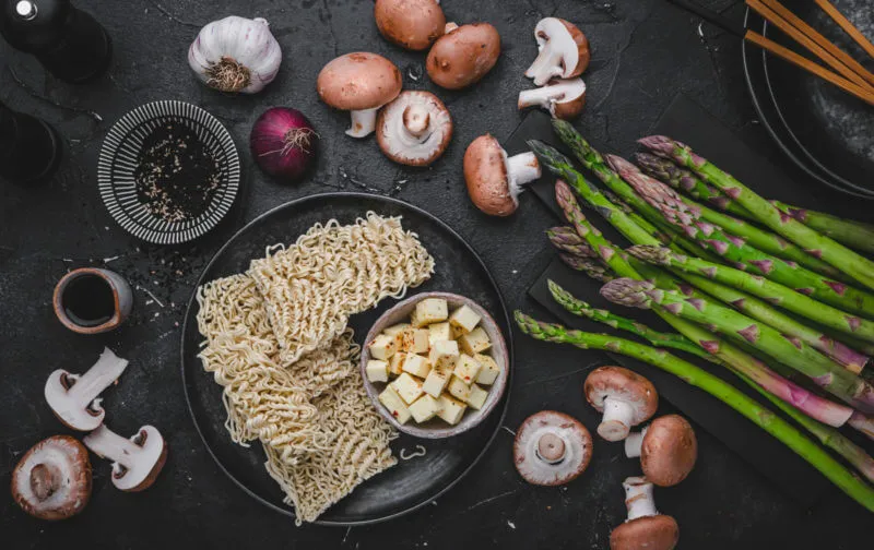 Ausgebreitete Zutaten für eine vegetarische Ramen Suppe in kleinen Schüsseln: Ramen-Nudeln, Halloumi, braune Champignons, Brühe und frischer grüner Spargel