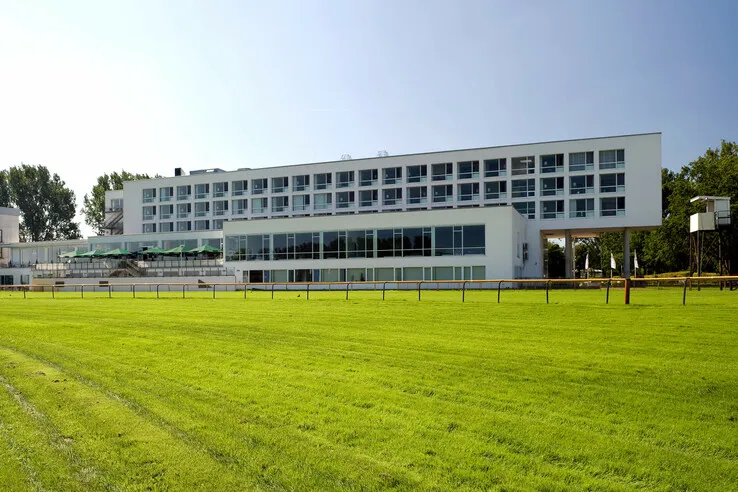 Außenansicht des ATLANTIC Hotel Galopprennbahn – ein modernes Hotelgebäude mit großen Fenstern und grünen Akzenten, umgeben von Bäumen und Rennbahn.