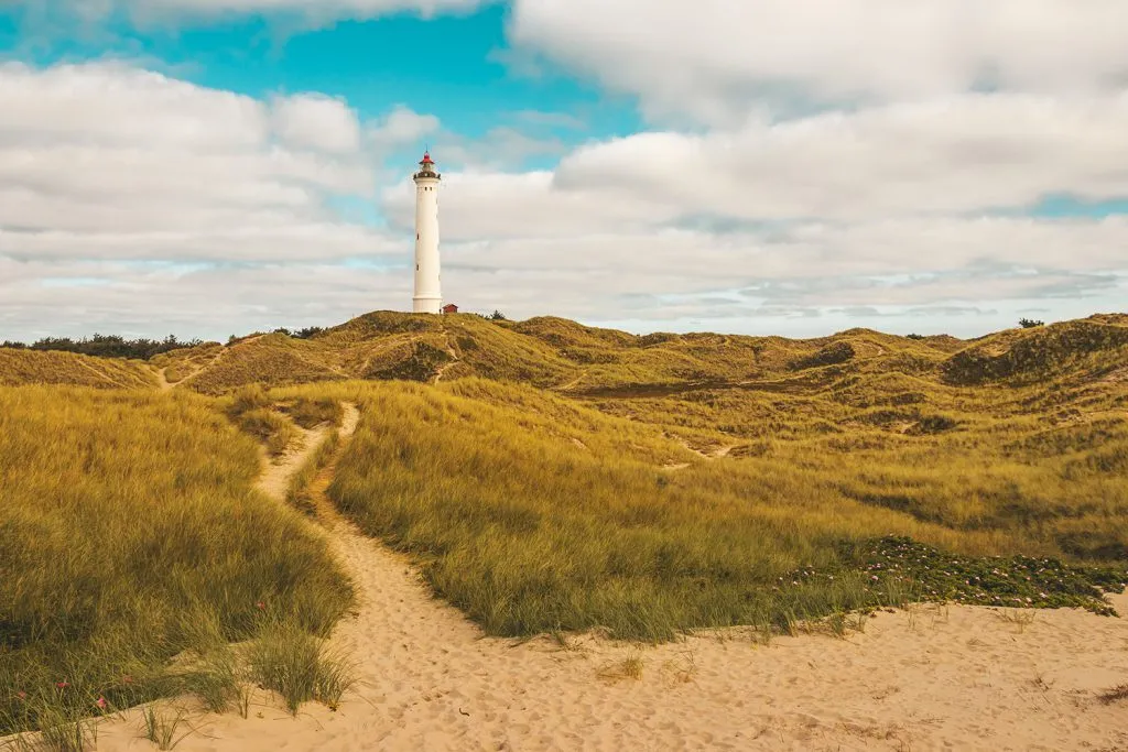 Aussenansicht des Lyngvig Fyr, ein roter Leuchtturm auf einer grünen Düne unter blauem Himmel