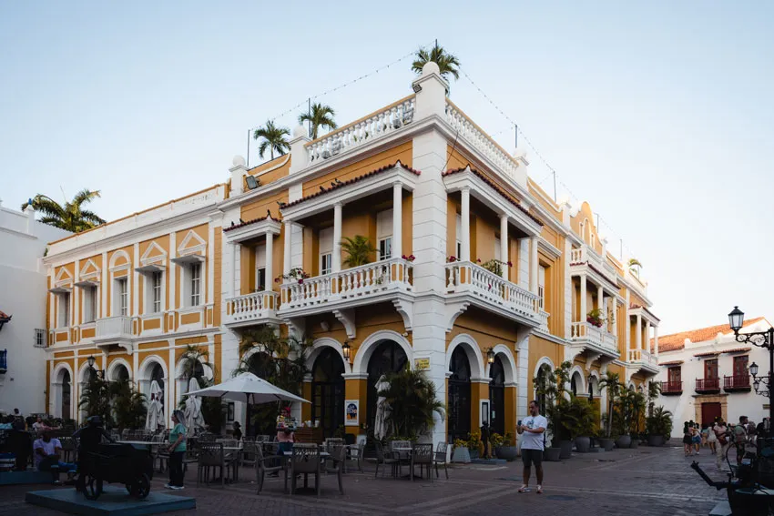 Belebter Platz mit einem historischen gelben Gebäude, Palmen und Balkonen, mit Außensitzplätzen und Besuchern