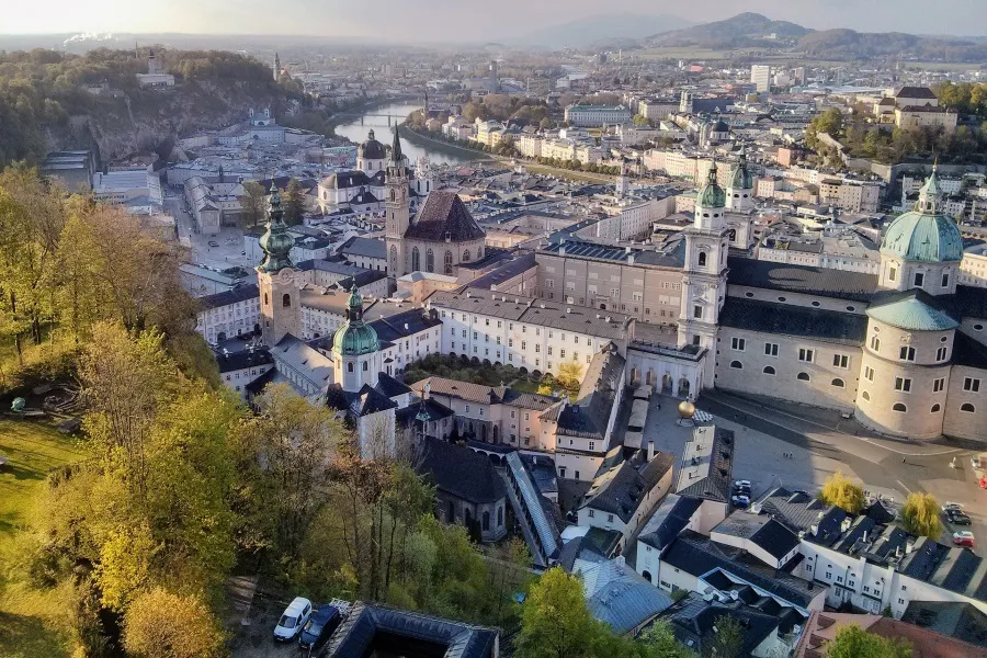 Blick auf Salzburg, Österreich, von der Festung Hohensalzburg. Die Stadt erstreckt sich mit ihren historischen Gebäuden, Kirchen und Flüssen unter einem bewölkten Himmel. Im Vordergrund sind Bäume und Grünflächen zu sehen.