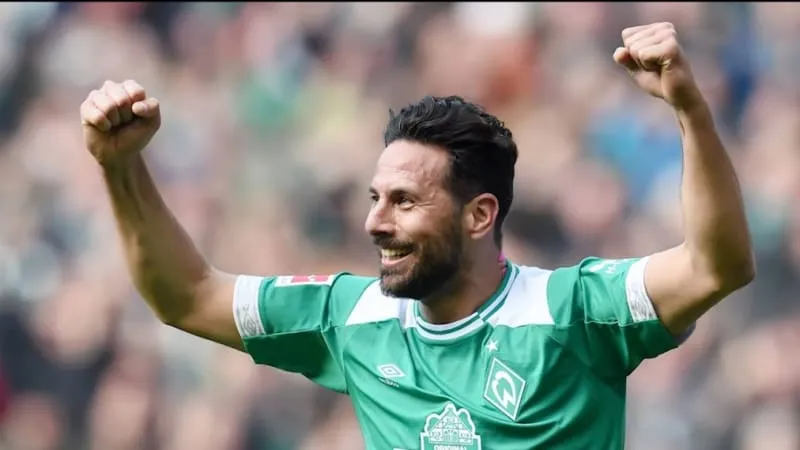 Claudio Pizarro erhält eine Auszeichnung als ältester Torschütze der Bundesliga und lächelt, im Hintergrund eine Leinwand mit seinem Namen und dem Logo von Werder Bremen.