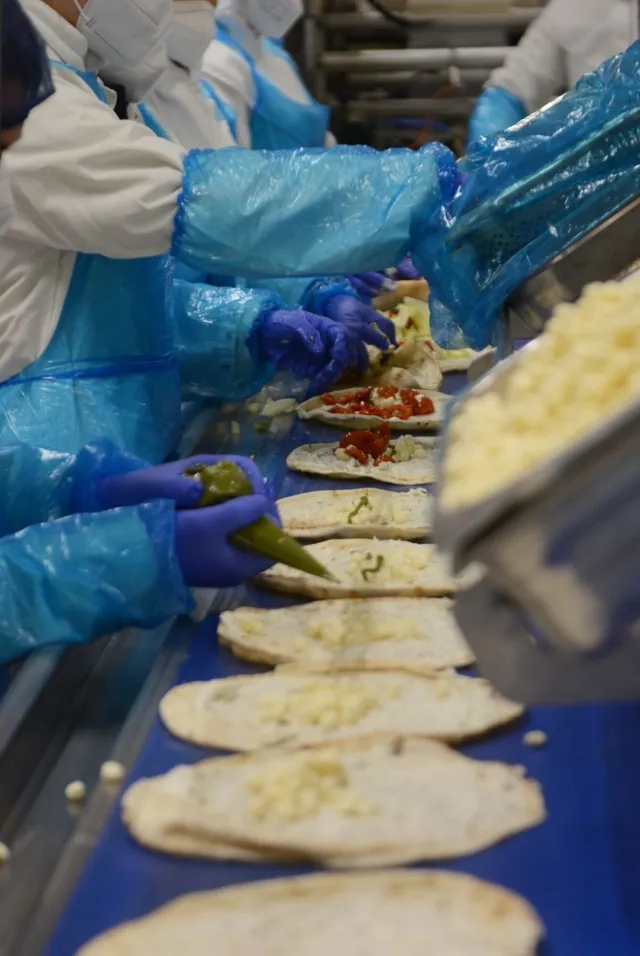 Das Bild zeigt LebensmitteltechnikerInnen in Schutzkleidung, die an einem Fließband Pizzen mit verschiedenen Zutaten belegen.