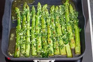 Das vorbereitete Olivenöl-Kräuter-Knoblauch-Gemisch wird über den bräunenden grünen Spargel in der Pfanne gegossen