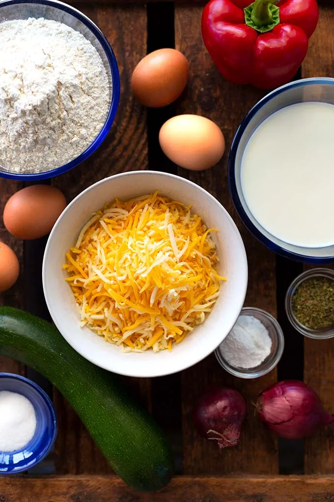Die Zutaten für die Gemüse-Quiche in Schüsseln und auf dunklem Holzuntergrund. Zu sehen sind ganze Eier, eine rote Paprika, Mehl, Milch, geriebener Käse, Salz, Kräuter, zwei ganze rote Zwiebeln und eine Zucchini.