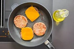 Drei Burger-Pattys braten in einer Pfanne, auf zwei davon liegt bereits eine Scheibe Cheddar-Käse, die langsam schmilzt.