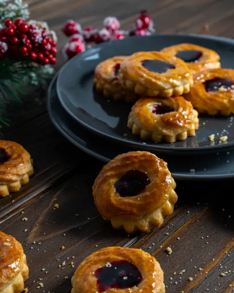 Drei gestapelte Pfauenaugen Gebäckstücke, eines davon angeschnitten, zeigen die feine Schichtung aus Mürbeteig, Makronen und Fruchtfüllung