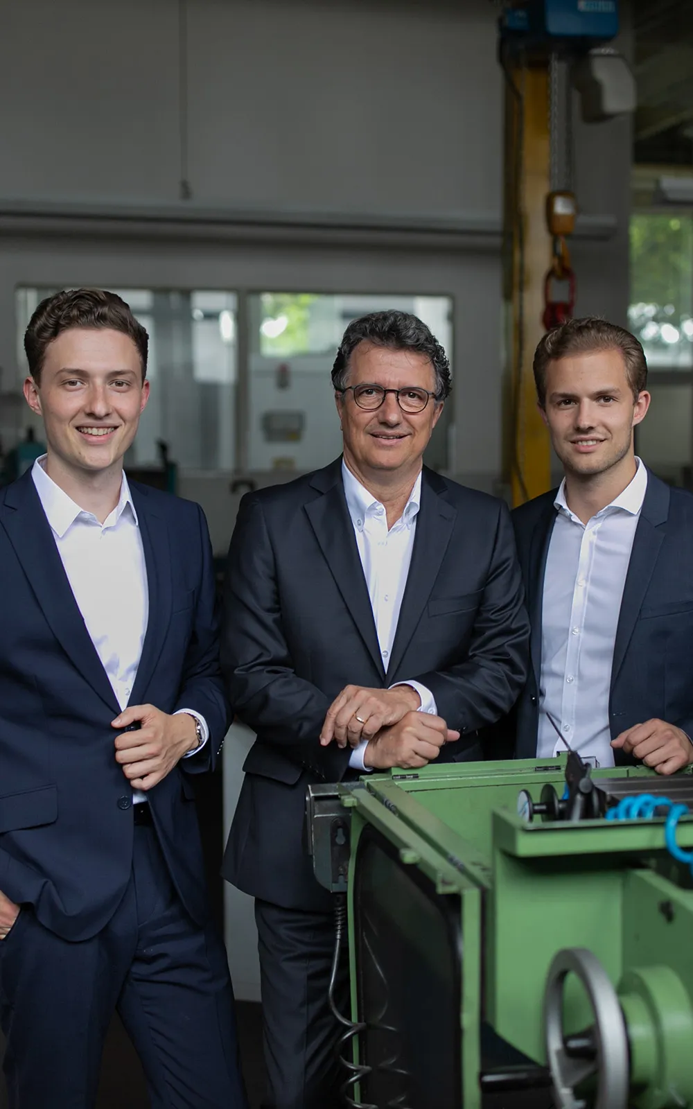 Drei Männer in Anzügen lächeln in einer Werkstatt in die Kamera