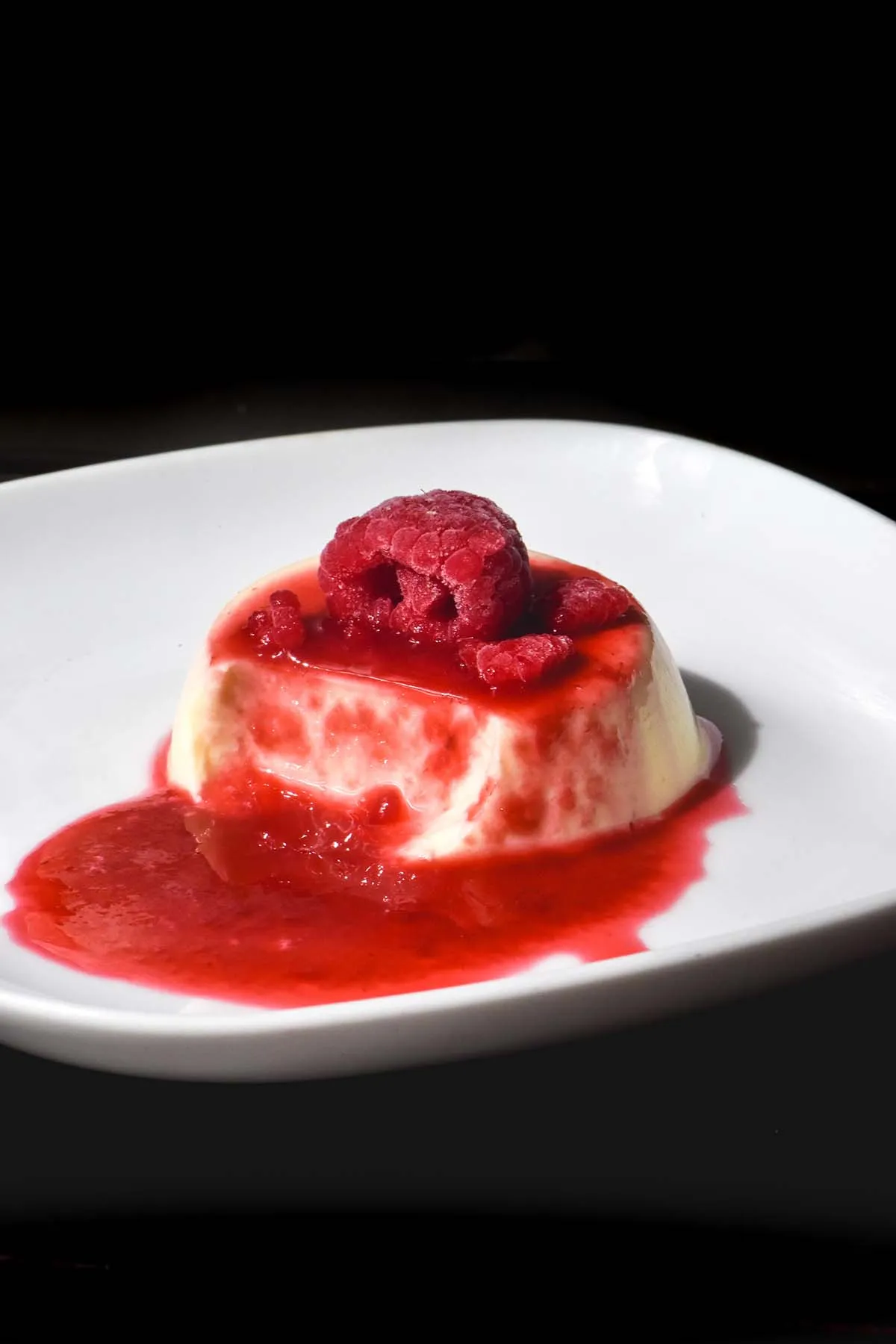 Dunkel inszenierte Panna Cotta mit reichlich Himbeercoulis und zusätzlichen Himbeeren auf einem kleinen weißen Teller.