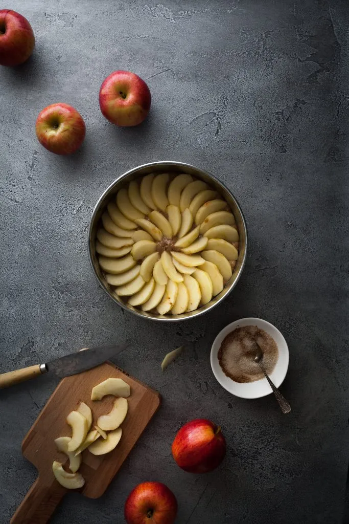 Ein Apfelkuchen in der Springform, umgeben von Zimtzucker und frischen Apfelscheiben, kurz vor dem Backen.