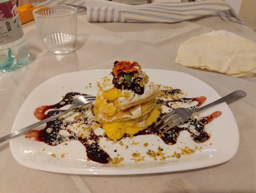 Ein dekonstruiertes Millefoglie-Dessert auf einem weißen Teller mit Schichten aus flockigem Gebäck und cremigem Pudding, garniert mit Orangenzesten, Beeren-Coulis und einer zarten essbaren Blume, die neben Besteck und einem Wasserglas auf den Genuss wartet.