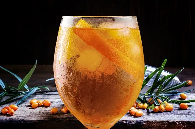Ein Glas Sanddorn Spritz auf einer hellen Fläche, dekoriert mit einer Orangenscheibe und Eiswürfeln, fängt das Licht ein und zeigt die leuchtend orange Farbe des Getränks.