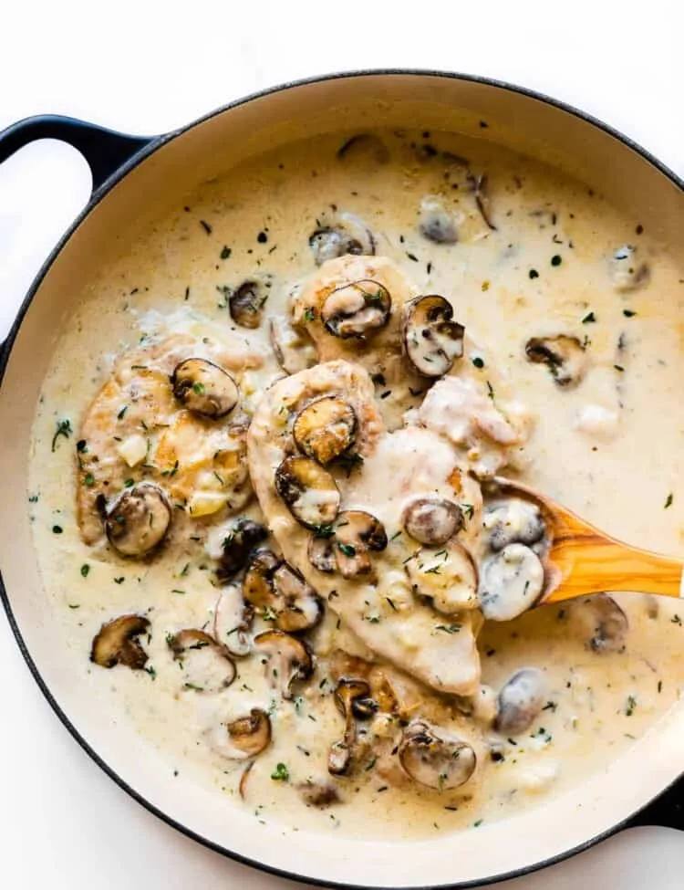 Ein Holzlöffel hebt Champagnerhähnchen mit einer cremigen Pilzsoße aus einer Pfanne,