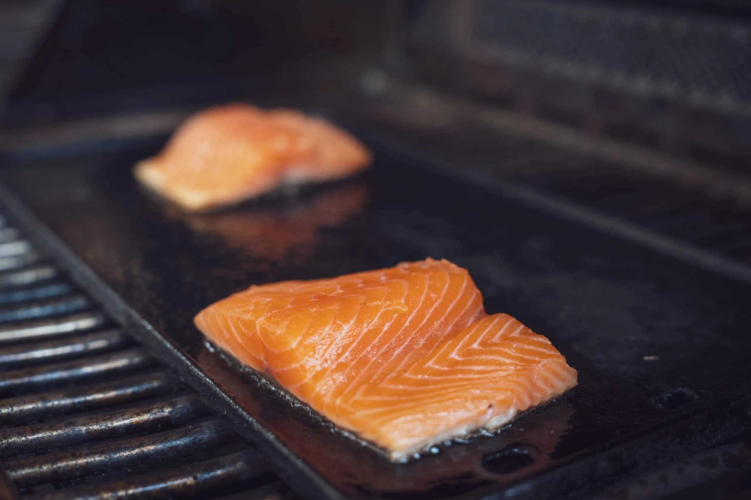 Ein Lachsfilet wird auf einer Plancha gegrillt, wodurch eine leckere Kruste entsteht, während das Innere saftig bleibt. Der Fisch ist leicht mit Kräutern und Gewürzen bestreut.