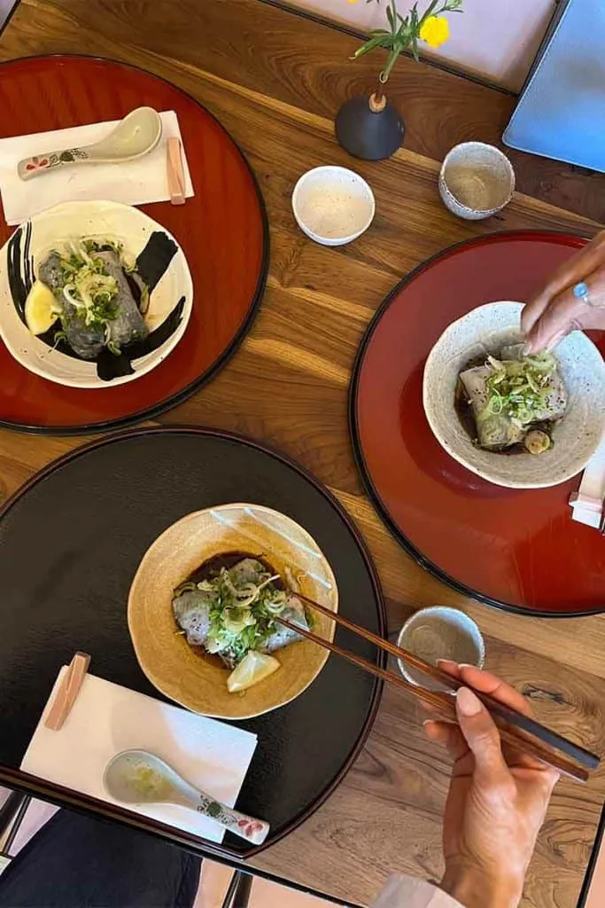 Ein liebevoll gedeckter Tisch im Pink Pig Japanese Cuisine in Zagreb mit drei Schalen eines delikaten Gerichts, wie Teigtaschen oder Fisch, in dunkler Soße, garniert mit Frühlingszwiebeln und Zitrone.