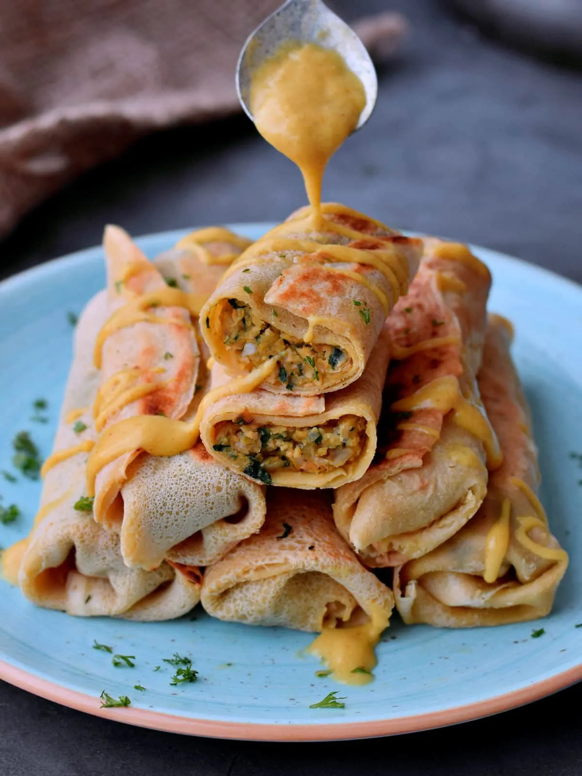 Ein Löffel träufelt vegane Käsesauce über eine Pyramide aus herzhaften Crêpes, gefüllt mit Kichererbsen, Champignons und Spinat.
