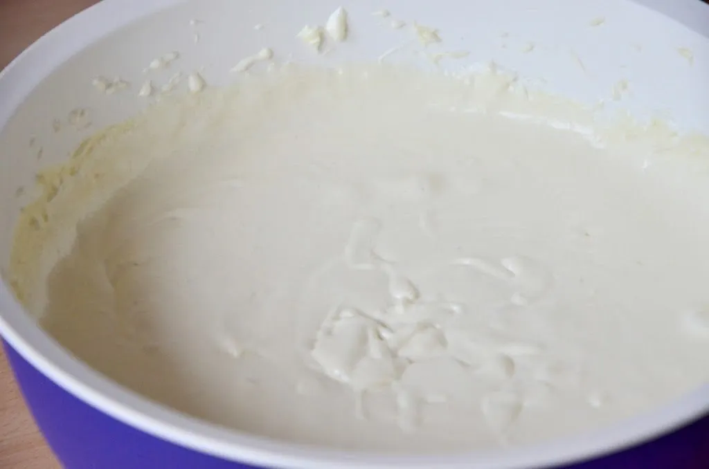 Ein Löffel voll geschmolzener weißer Schokolade wird von einer Glasschüssel in eine andere Rührschüssel mit Frischkäse gegeben