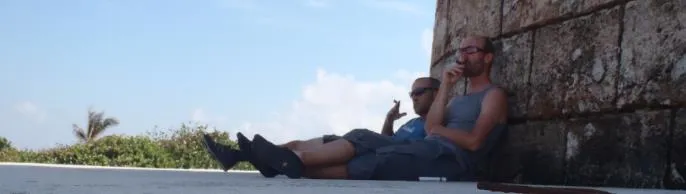 Ein Mann sitzt vor einem farbenfrohen Gebäude in Havanna und raucht eine Zigarre.