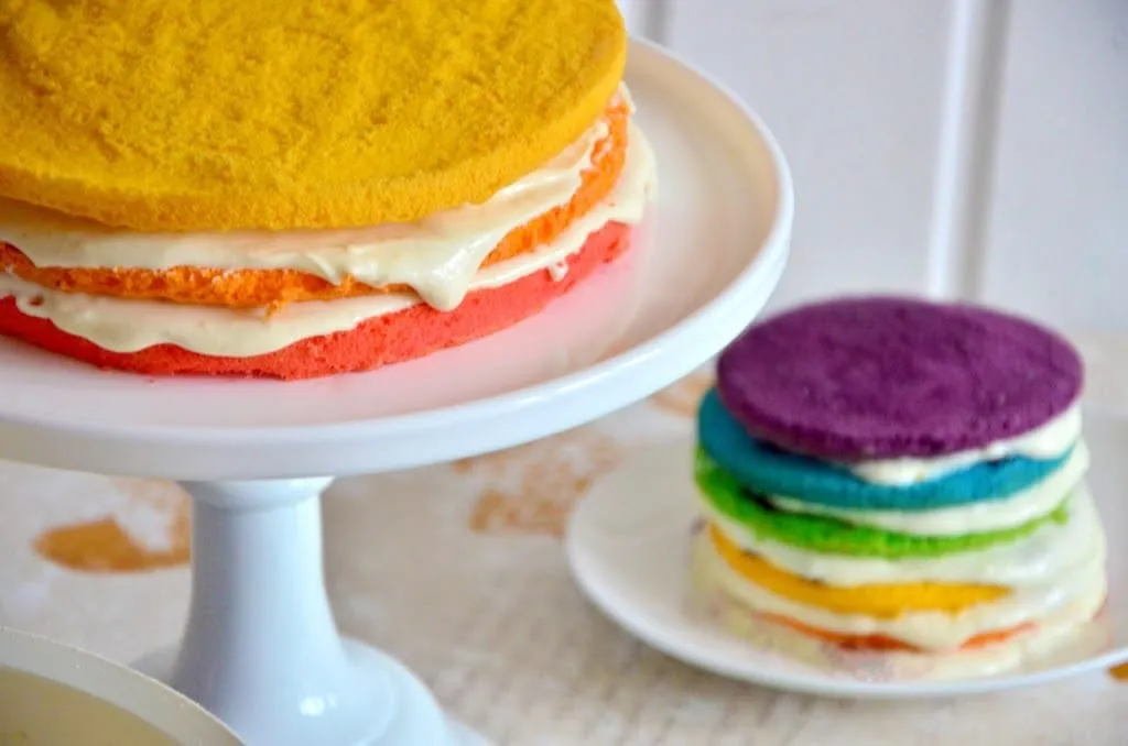 Ein orangefarbener Biskuitboden wird vorsichtig auf die weiße Frischkäse-Schicht des roten Bodens gelegt