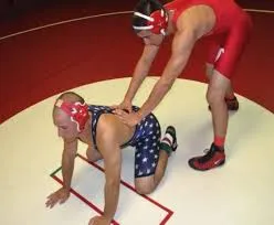 Ein Ringer in rotem Anzug führt einen optionalen Start aus, während sein Gegner in der Bodenlage kniet, eine typische Szene im Ringkampf.