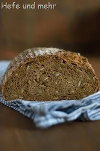 Ein rustikales, dunkles Roggenbrot mit Kürbiskernen und Sonnenblumenkernen, frisch aus dem Ofen, steht auf einem Schneidebrett.