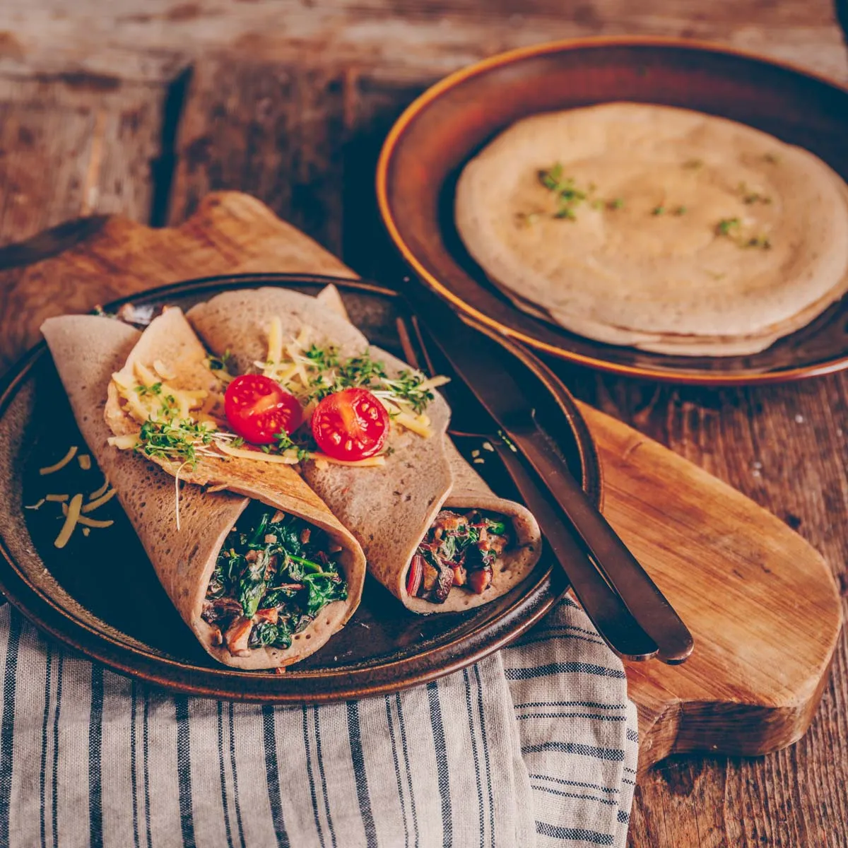 Ein Stapel goldbrauner Buchweizenpfannkuchen mit einer cremigen Spinatfüllung, garniert mit frischen Kräutern, die auf einem rustikalen Holztisch serviert werden.