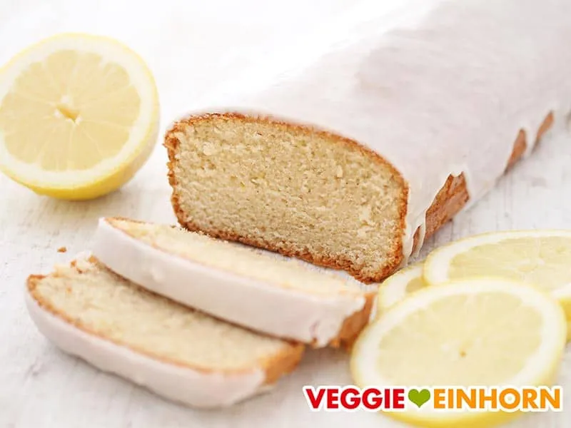 Ein Stück veganer Zitronenkuchen mit frischer Zitronenglasur auf einem Teller, umgeben von Zitronenscheiben.