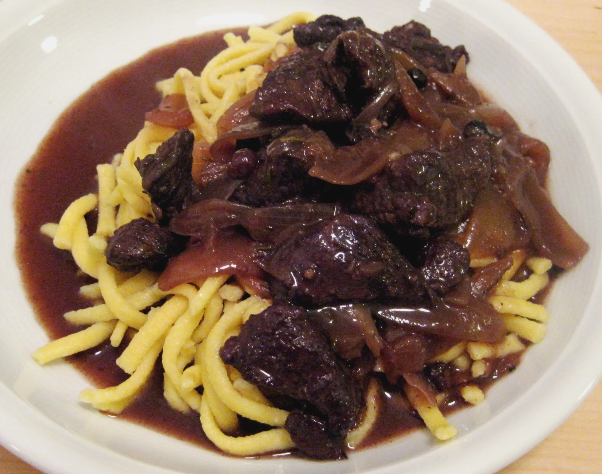 Ein Teller dampfender Hirschgulasch, garniert mit Preiselbeeren und Petersilie, steht auf einem rustikalen Holztisch. Im Hintergrund ist eine Flasche Rotwein zu sehen.