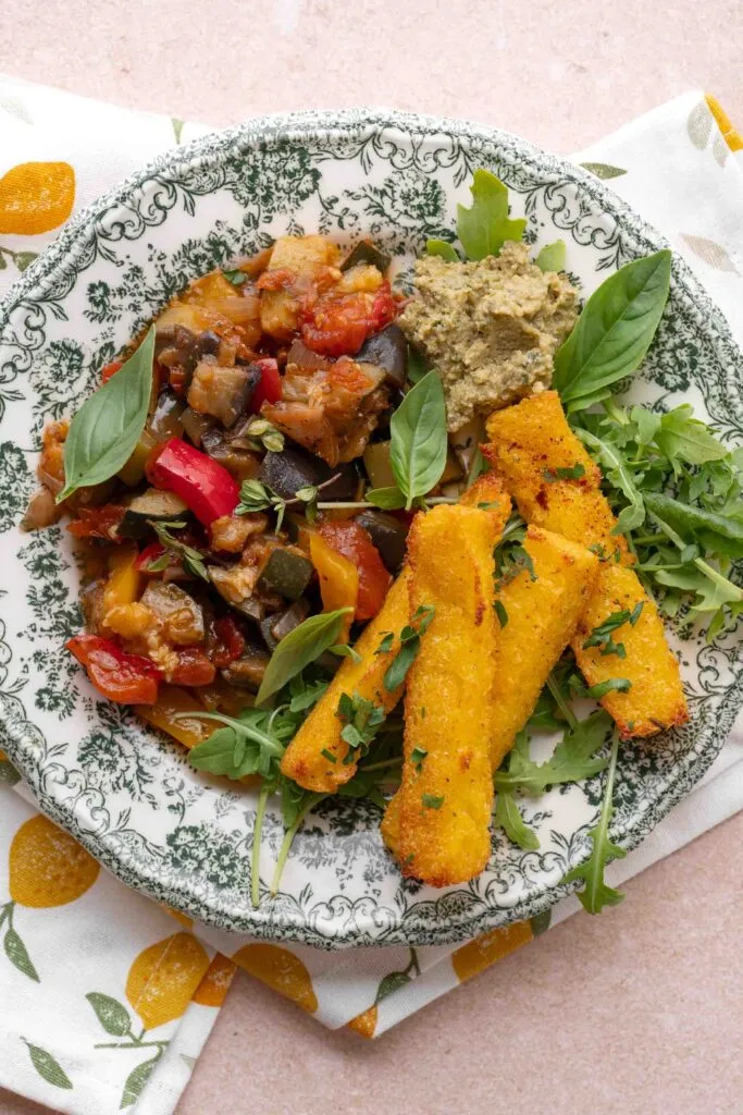 Ein Teller mit Polenta-Chips, Gemüse-Ratatouille, einer Portion grünem Dip und frischen Basilikumblättern, angerichtet auf einer gemusterten Platte.