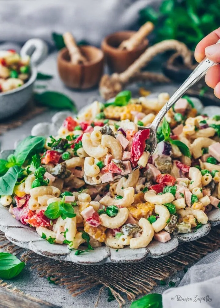 Ein Teller mit veganem Nudelsalat, garniert mit veganer Fleischwurst, Mayonnaise und verschiedenen Gemüsesorten, präsentiert als eine gesunde und ansprechende Mahlzeit.