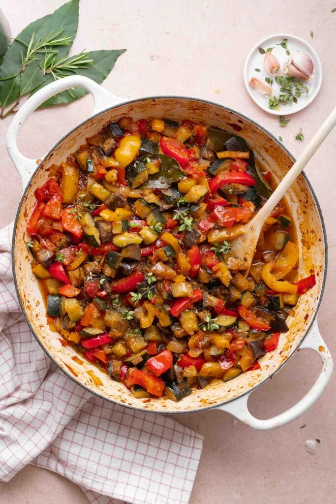 Ein weißer Topf mit gekochtem Ratatouille, das aus gehackten Zucchini, Auberginen, Paprika und Tomaten besteht, steht auf einer karierten Serviette mit einem Holzlöffel und frischen Kräutern in der Nähe.
