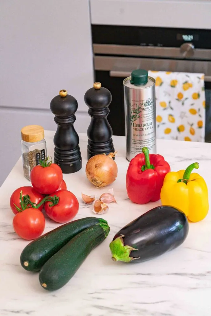 Eine Auswahl an frischem Gemüse, Pfeffermühlen, Olivenöl und Gewürzen sind auf einer Küchentheke vor einem Ofen arrangiert.