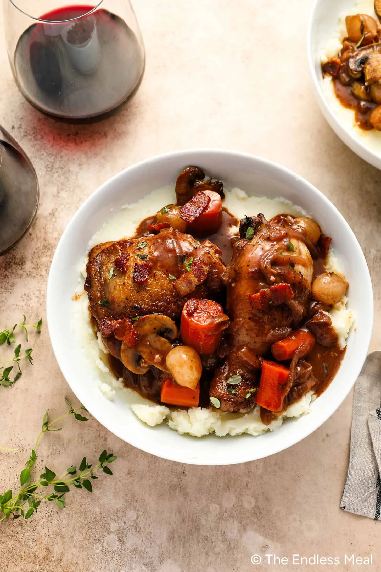 Eine dampfende Schüssel Coq au Vin mit zartem Hähnchen, Karotten, Pilzen und Zwiebeln auf butterartigem Kartoffelpüree steht einladend auf dem Tisch neben zwei Gläsern Rotwein.