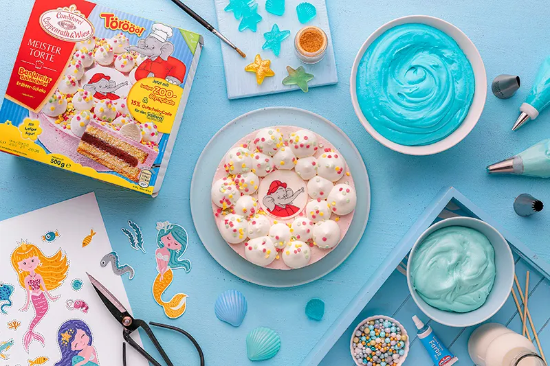 Eine farbenprächtige Meerjungfrau Torte, verziert mit einem Meerjungfrauen-Caketopper, süßen Muscheln und glitzernden Zuckerperlen, perfekt für einen Kindergeburtstag.