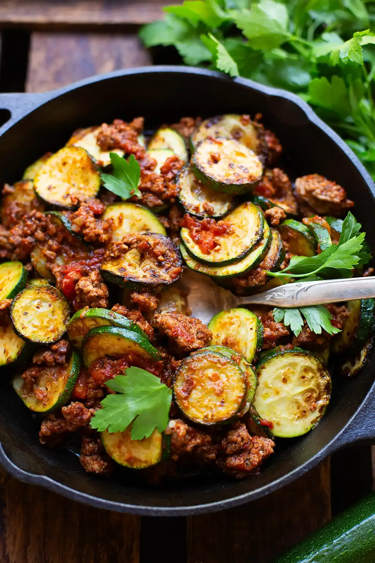 Eine fertige Zucchini-Hackfleisch-Pfanne in einer schwarzen Pfanne auf einem dunklen Holzuntergrund, garniert mit glatter Petersilie. Ein Bund frische Petersilie liegt neben der Pfanne. Eine Gabel steckt in der Low-Carb-Zucchini-Pfanne. Das Gericht bietet eine ausgewogene Mahlzeit.