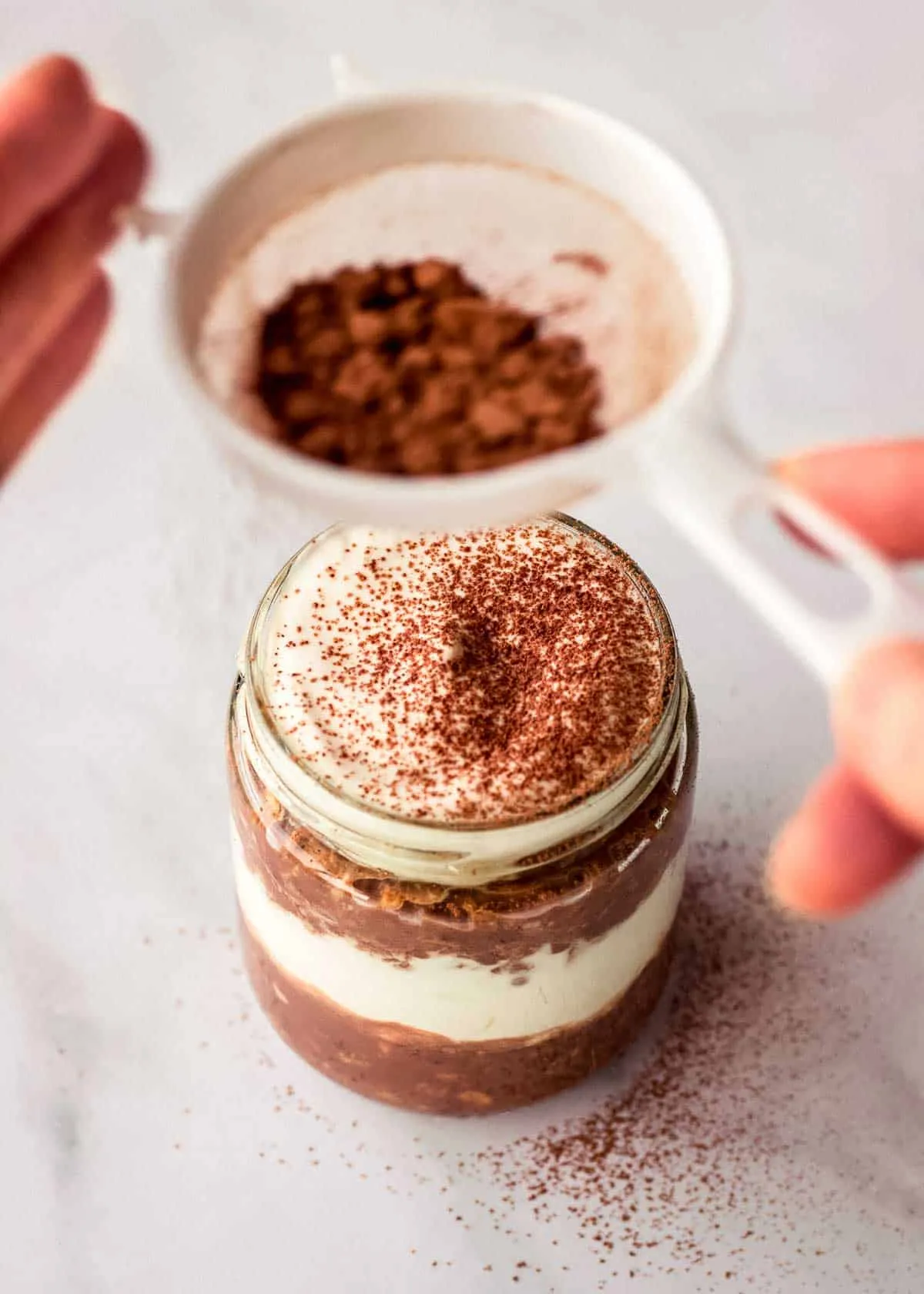Eine Frau siebt Kakaopulver durch ein Sieb über ein Glas mit geschichteten Tiramisu Overnight Oats.