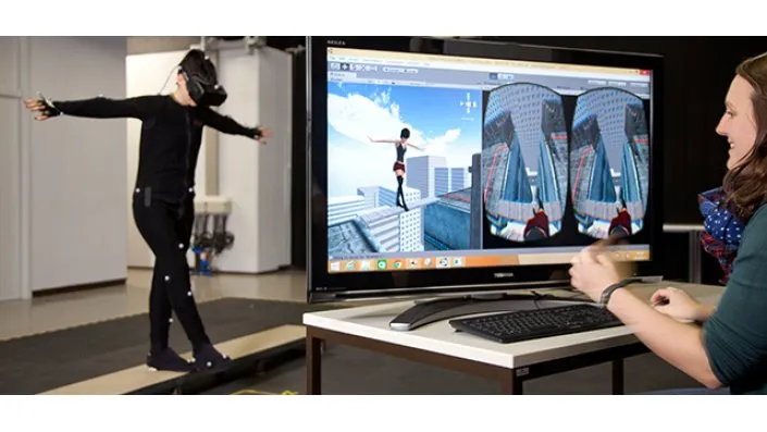 Eine Frau trägt eine VR-Brille und balanciert auf einer Planke, während eine andere Person ihre Erfahrung auf einem Monitor verfolgt.