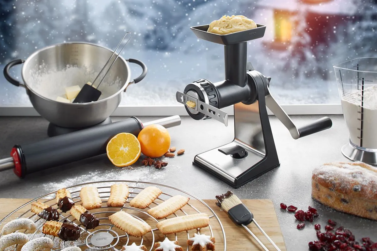 Eine Gebäckpresse fertigt winterliche Plätzchen, begleitet von Orangen und Backzutaten auf einer weihnachtlichen Küchentheke.