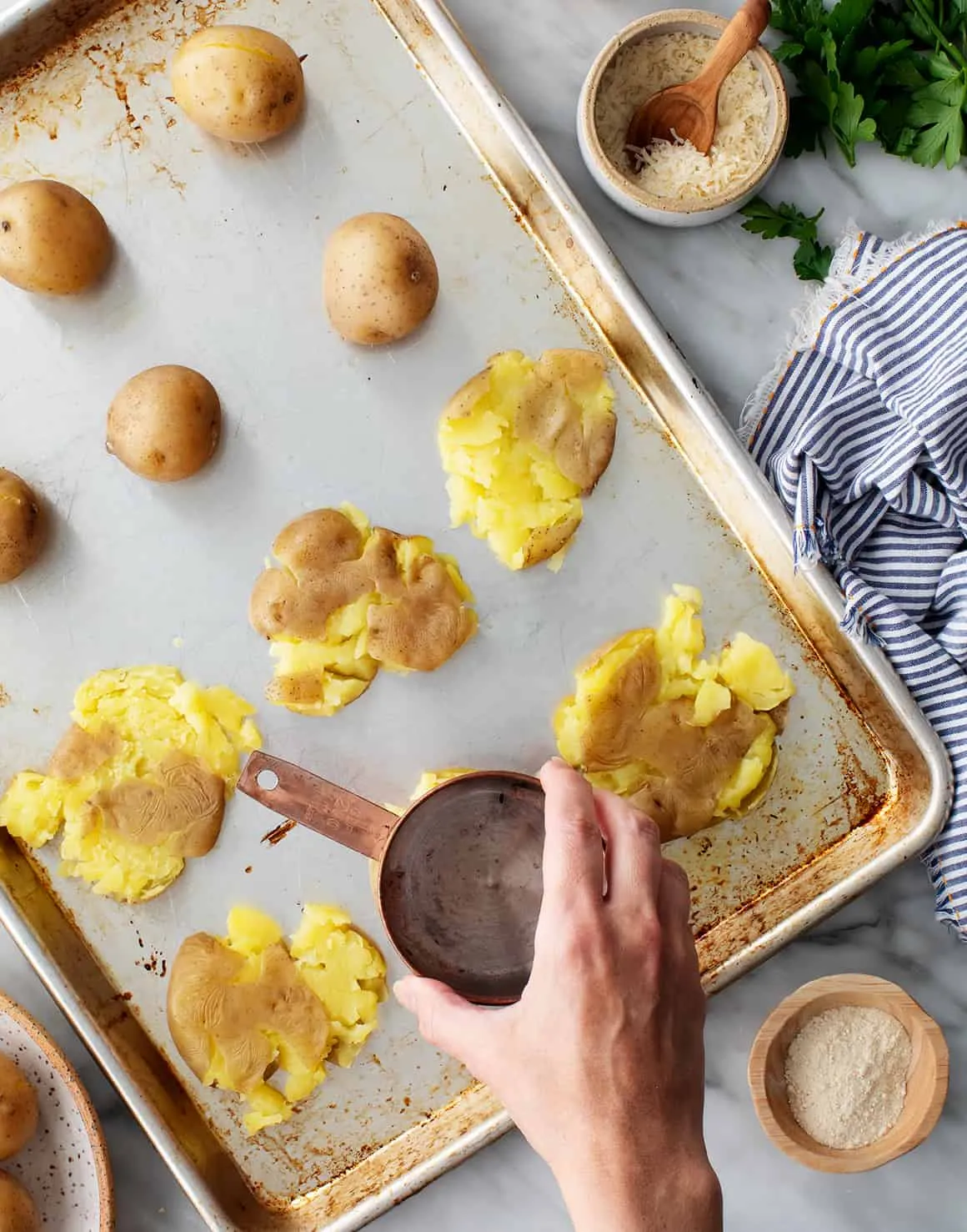 Eine gekochte Kartoffel wird mit dem Boden eines Messbechers auf einem geölten Backblech zerdrückt