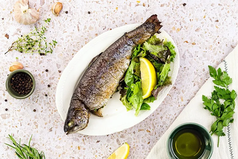 Eine goldbraun gebackene Forelle auf einem Backblech, umgeben von frischen Kräutern und Zitronenscheiben, ideal für fettarme Fischrezepte
