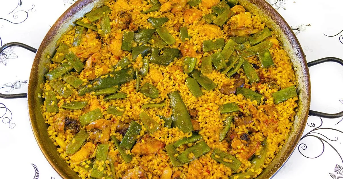 Eine goldene Paella Valenciana mit Reis, Hähnchen, Kaninchen und Gemüse, garniert mit Zitronenspalten, serviert in einer großen, flachen Paellapfanne auf einem rustikalen Holztisch
