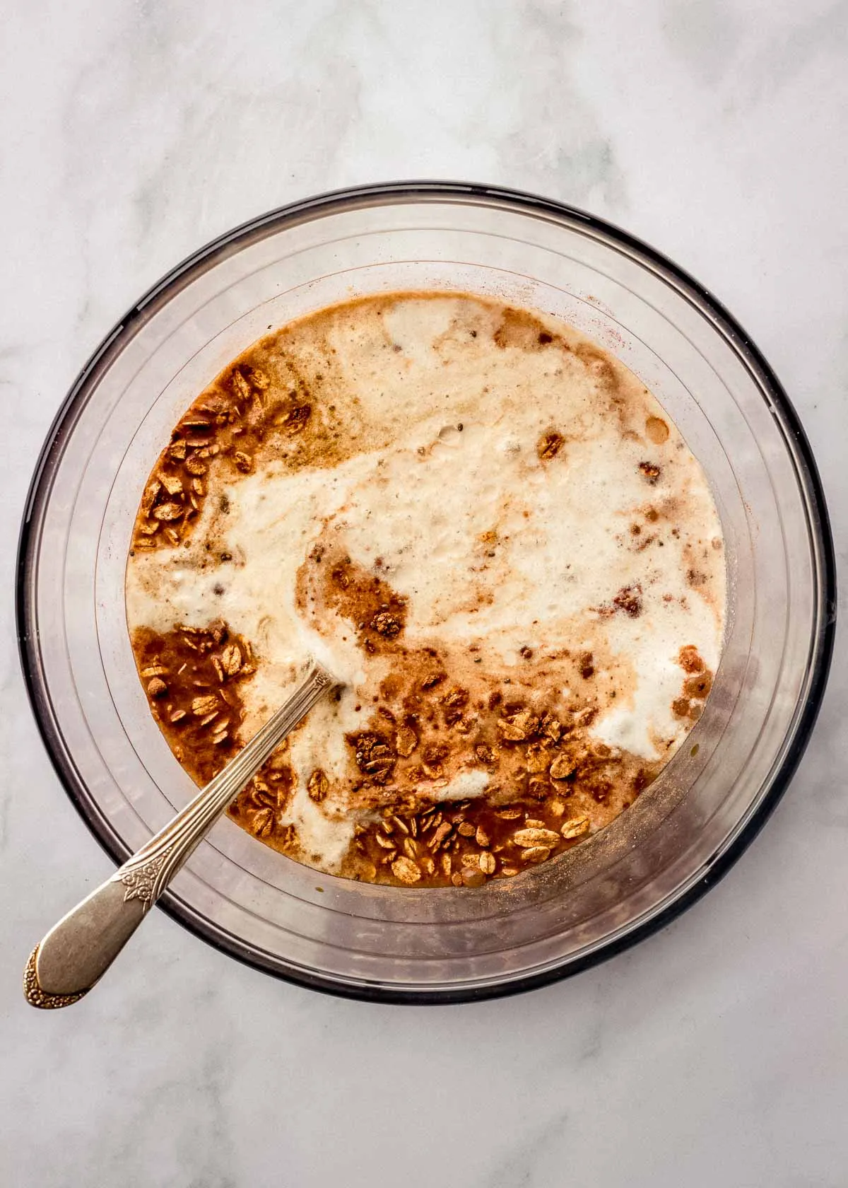 Eine große Glasschüssel mit Tiramisu Overnight Oats Zutaten mit einem Löffel darin, bereit zum Umrühren.