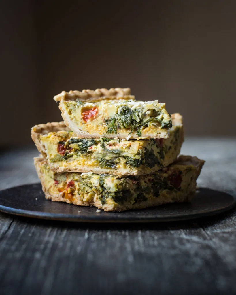 Eine halbe vegane Quiche mit Spinat, Dill und Tomaten, angerichtet auf einem rustikalen Holzbrett.