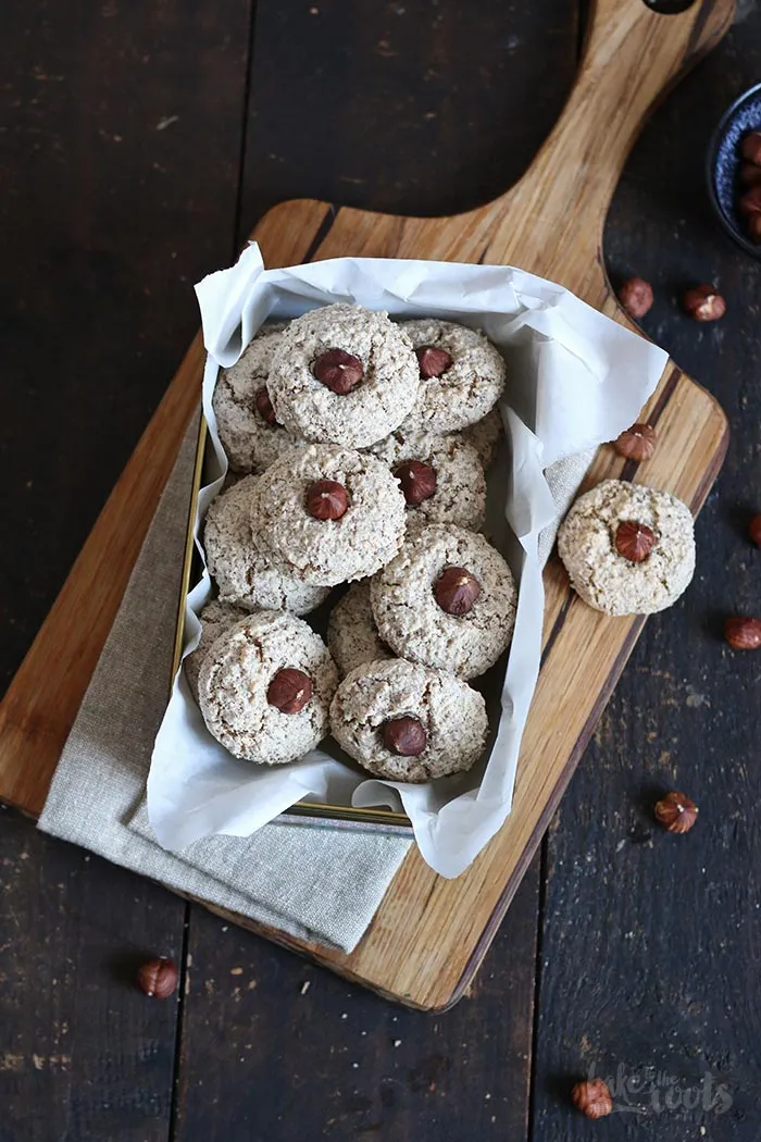 Eine Hand drückt leicht auf die Oberfläche einer Haselnussmakrone, um die Konsistenz zu prüfen. Die Makrone ist goldbraun und hat eine leicht rissige Oberfläche, mit einer ganzen Haselnuss in der Mitte.