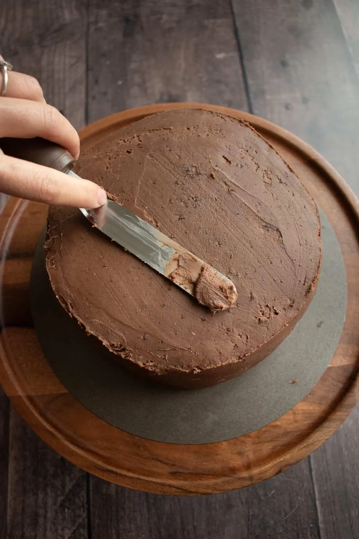 Eine Hand glättet Schokoladenfrosting auf der Oberseite eines runden Kuchens.
